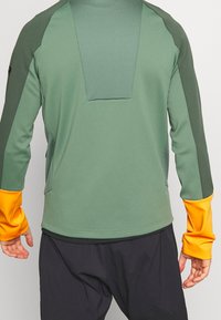 Groene sportieve lange mouwen top met een mesh textuur op de achterkant en oranje accenten op de mouwen. Gladde stof, aangesloten ontwerp.