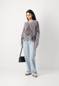 Grau strukturierter gestrickter Pullover mit braunem Muster, kombiniert mit hellen Jeans. Das Modell hält eine kleine schwarze Handtasche. Lockere Schuhe sind sichtbar.