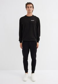 Sudadera negra con el texto "INSIDE", mangas raglán, combinada con pantalones deportivos negros que tienen bolsillos y dobladillos ajustados, llevada con zapatillas blancas.