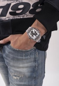 Orologio quadrato, trasparente, con accenti argentati, quadrante bianco e lancette nere. Indossato su un polso, abbinato a jeans chiari sfilacciati.