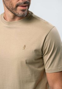 Mees hele näokarvaga, kandes beežikat lühikese varrukaga T-särki, millel rinnal on väike tikitud logo.