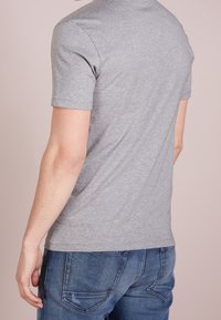 Šedé bavlněné tričko s krátkým rukávem a kulatým výstřihem, viděné ze zadní strany, v kombinaci s modrými džíny. Viditelné švy.