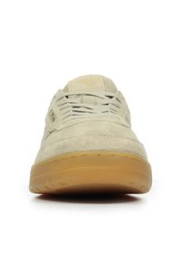 Zapatilla de ante beige con puntera redonda, cordones beige y suela de goma. Presenta una etiqueta con el logo en el lateral y un acabado texturizado en la parte superior.