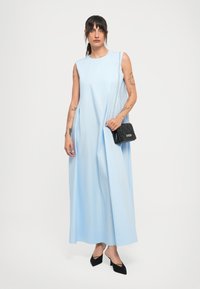 Donna con tatuaggi che indossa un abito maxi senza maniche di un blu chiaro, tacchi neri e porta una piccola borsa a spalla nera trapuntata.