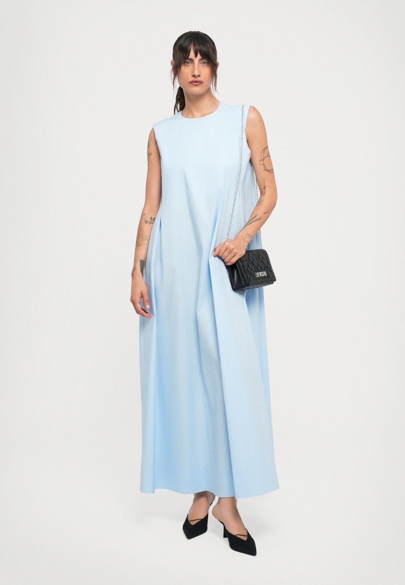 Donna con tatuaggi che indossa un abito maxi senza maniche di un blu chiaro, tacchi neri e porta una piccola borsa a spalla nera trapuntata.