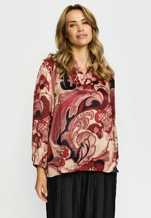 Kvinde med blondt hår iført en løs, langærmet bluse med røde, lyserøde og sorte blomster-swirls samt et sort plisseret nederdel.