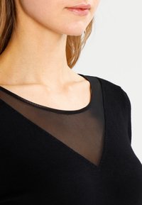 Haut noir avec un design en V et un panneau en maille transparent au-dessus de l'encolure. Texture lisse, forme ajustée et style minimaliste.