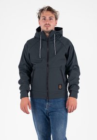 Uomo con capelli biondi disordinati, che indossa una giacca a zip nera con cappuccio e jeans blu, in piedi davanti a uno sfondo bianco uniforme.