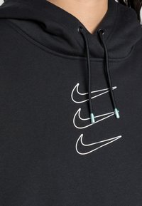 Svart hoodie i en blandning av bomull med fyra vita Nike-logotyper på framsidan. Dragsnören har gröna spetsar.