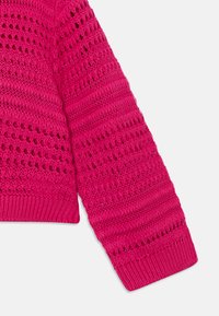 Lys rosa strikket genser med en teksturert design, som har hullmønster og ribbestrikkede mansjetter. Stoffet ser lett og pustende ut.
