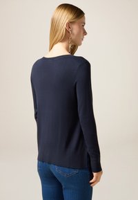 Maglione a maglia blu navy con scollatura a girocollo, maniche lunghe, spacchi laterali e vestibilità rilassata. Texture morbida, design casual. Visibili jeans blu denim.