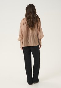 Blusa in seta beige chiaro con maniche a sbuffo, dettagli arricciati sulle spalle e bottoni decorativi sui polsini. Abbinata a pantaloni neri.