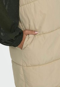 Beige Puffer-Weste mit einer Seitentasche, die über eine glatte Textur und ein dezentes Steppmuster verfügt. Die Überlagerung umfasst einen dunkelgrünen Ärmel mit Punktmuster.