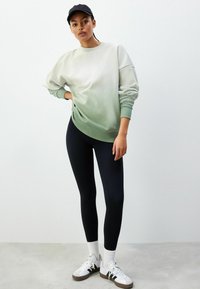 Pulover cu gradient de la verde deschis la alb, cu umerii lăsați, asortat cu colanți negri și adidași albi. Prezentat pe un fundal neutru.