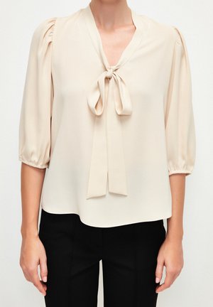 Femme portant un chemisier crème avec des manches bouffantes jusqu'au coude et un nœud à l'encolure, associé à un pantalon noir.
