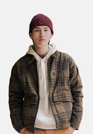 Giovane uomo che indossa un berretto bordeaux, felpa beige e giacca a quadretti marroni, in piedi con le mani in tasca contro uno sfondo neutro.
