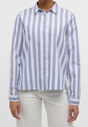 Kvinde iført en hvid skjorte med knapper med lodrette mørkeblå striber og hvide bukser, hænderne afslappet ved siderne.