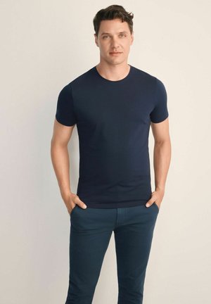 Mężczyzna stoi z rękami w kieszeniach, ubrany w dopasowany granatowy t-shirt i ciemnoniebieskie spodnie na jednolitym jasnym tle.