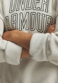Svetlošedý mikina s čiernym nápisom "UNDER ARMOUR". Má jemnú textúru a široké manžety. Dizajn obsahuje rebrové detaily na rukávoch.