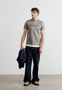 Νεαρός άνδρας με γκρι μπλούζα με σχέδια πάνω από λευκό πουκάμισο με μακριά μανίκια, σκούρα τζιν με φαρδιά πόδια, κρατώντας σκούρο σακάκι, στέκεται σε γκρι δάπεδο.