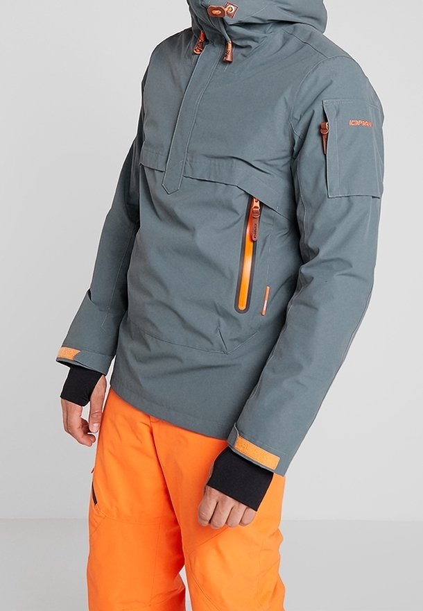 Homme portant une veste grise avec des fermetures éclair orange et un pantalon orange vif, se tenant devant un fond clair uni.