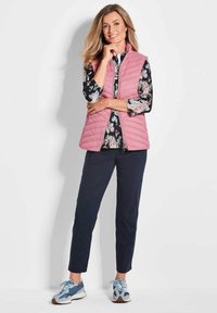 Rosa gesteppte Weste über einem gemusterten schwarzen Hemd, dunkle Hose und blaue Turnschuhe. Glatte Textur mit einem Frontreißverschluss und Seitentaschen.