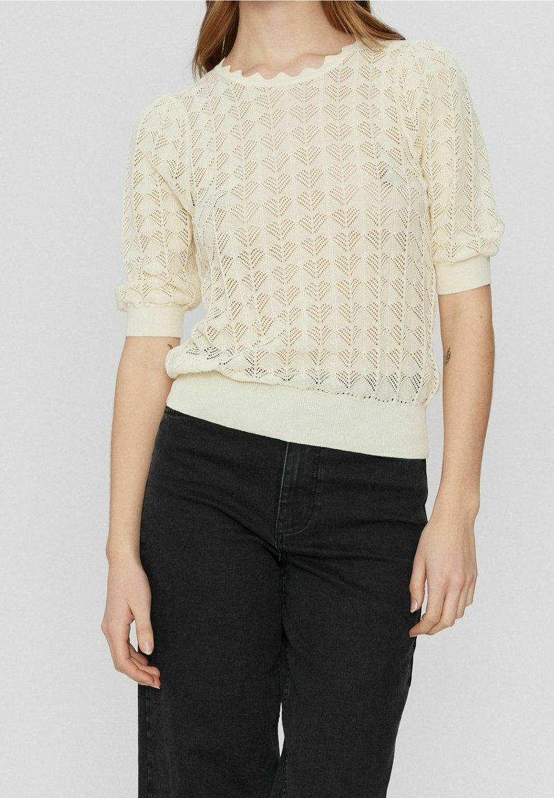 Pletený svetr v krémové barvě s texturovaným listovým vzorem, vlnkovým výstřihem, krátkými puff rukávy a žebrovaným lemem, kombinovaný s tmavými džíny.