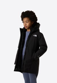 Μαύρο μονωμένο παρκά με κουκούλα από ψεύτικη γούνα, εμπρόσθιο φερμουάρ, πλαϊνές τσέπες και λογότυπο στο στήθος. Το εσωτερικό φόδρα φαίνεται μέσα από το ανοιχτό φερμουάρ.