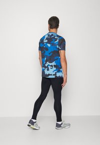 Camisa de manga corta de camuflaje azul, leggings ajustados negros y zapatillas grises con detalles en amarillo, vistas desde atrás.