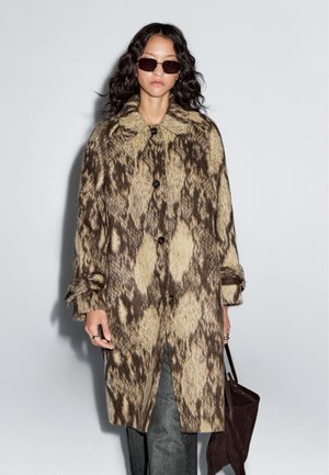 Knie-langer Mantel mit einem braun-beigen Tiermuster, aus strukturiertem Stoff, mit einem großen Kragen und einer Knopfleiste, kombiniert mit grauen Hosen.