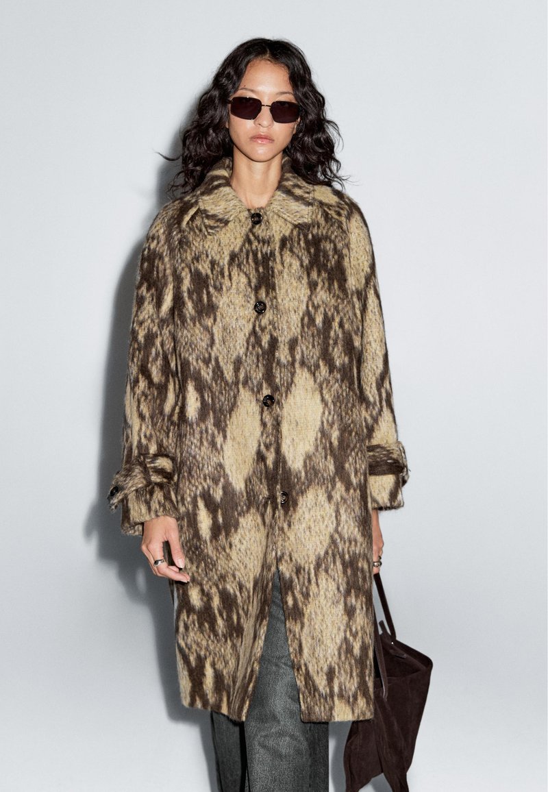 Knie-langer Mantel mit einem braun-beigen Tiermuster, aus strukturiertem Stoff, mit einem großen Kragen und einer Knopfleiste, kombiniert mit grauen Hosen.