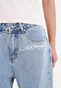 Light blue denim jeans feature a high waist design with a silver button and an embroidered "Ed Hardy" logo in white on the front pocket.