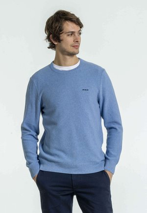 Pull en tricot bleu clair avec col et poignets côtelés, arborant un petit logo noir sur la poitrine. Porté par-dessus un t-shirt blanc.