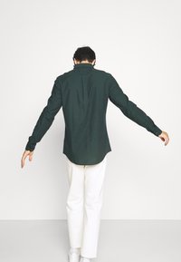 Camicia verde scuro con maniche lunghe, colletto e piega posteriore, abbinata a pantaloni bianchi e sneakers.