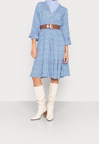 Modré šaty s květinovým vzorem, dlouhými rukávy a volánkovým lemem, stažené v pase širokým hnědým opaskem. Pár bílých vysokých bot.