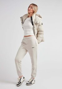 Pufjakke i lys beige med sorte detaljer, parret med cremede sweatpants med "JACKIT" logo, og sort-hvide sneakers.