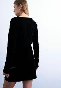 Femme aux cheveux châtain clair portant des boucles d'oreilles argentées, un haut à manches longues noir et une jupe noire, debout devant un fond blanc uni.