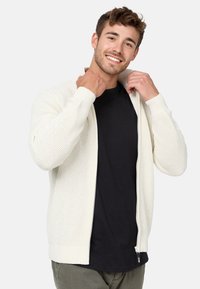 Hvid strikket zip cardigan med et tekstureret mønster, ribbede manchetter og kant. Bæret over en sort skjorte, justerer modellen kraven.