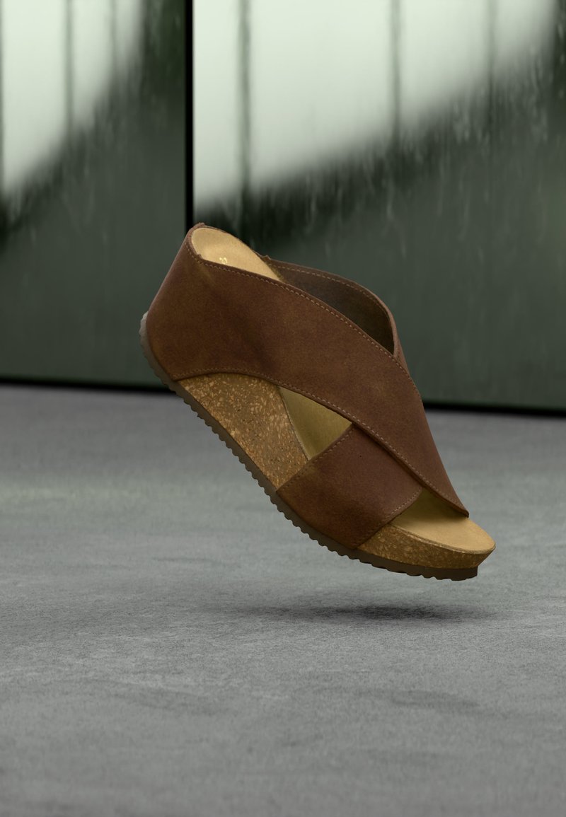 Καφέ πλατφόρμα σανδάλι με φελλό στη σόλα και λουριά που διασταυρώνονται, εμφανίζεται να αιωρείται πάνω από γκρίζα επιφάνεια με θολό φόντο.