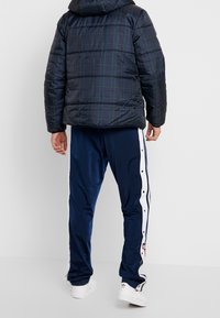 Navy puffer jacket with a plaid pattern, featuring a hood. Paired with matching pants, accented by white side stripes and snap buttons.