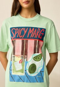 Ljust grön bomulls-t-shirt med en färgglad grafik av ett cocktailglas, avokado och limeklyftor mot en blå bakgrund.