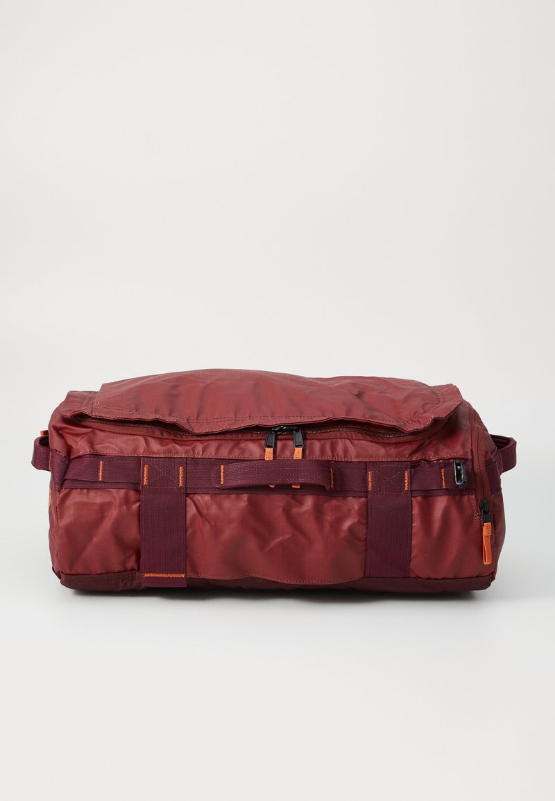 Sac de voyage en tissu rouge avec un dessus zippé, quincaillerie noire, accents orange et une sangle épaisse. Forme rectangulaire avec une texture lisse.