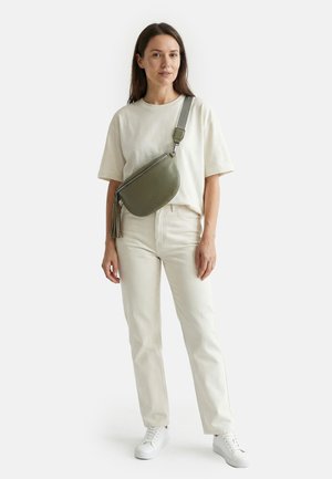 Woman standing wearing cream t-shirt and pants, white sneakers, and an olive green crossbody bag with wide strap and tassel detail.