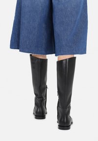 Black leather knee-high boots with a smooth texture, featuring side zippers and a rounded toe, paired with a blue denim skirt.