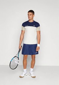 Sportininkas, vilkintis mėlynai baltą teniso aprangą, laiko raketę, stovėdamas prieš baltą fono, apsirengęs baltais sportiniais bateliais ir kojinėmis.