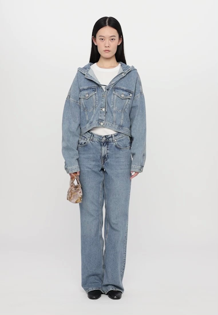 Veste en denim cropped bleu clair avec capuche, associée à un jean droit assorti. Le modèle tient un petit sac à main à motifs et porte des chaussures noires.