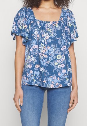 Nainen pukeutunut siniseen kukkakuosiseen lyhythihaiseen puseroon, jossa on neliskulmainen pääntie, ja vaaleansinisiin farkkuihin, seisoo yksiväristä taustaa vasten.