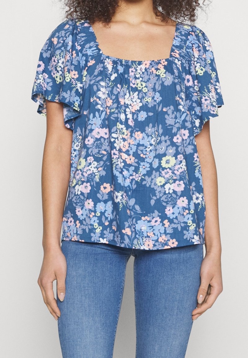 Femme portant un chemisier bleu à fleurs à manches courtes avec un col carré et un jean bleu clair, debout devant un fond uni.