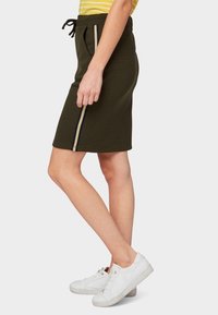 Sukně olivové zelené barvy, sahající po kolena, vyrobená z měkké tkaniny s páskem na stažení v pase a kontrastními bočními pruhy v žluté a námořnické modři. S kombinací s bílými teniskami.