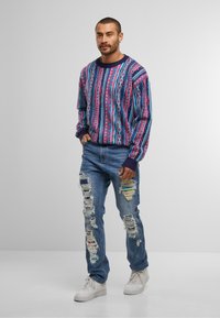 Bunt gestrickter Pullover mit geometrischen Mustern, gerippten Bündchen und Rundhalsausschnitt. Ausgefranste blaue Jeans mit mehreren Rissen und weißen Sneakern.